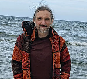 Foto unseres Referenten Steve Heitzer: Er lacht in die Kamera, hat braunes Haar, dass er zusammengebunden hat,und trägt ein blau-weiß gestreiftes Hemd