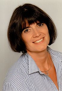 Foto unserer Referentin Szilvia Jördens: Sie lächelt, hat braunes schulterlanges Haar mit Pony und trägt eine blau-weiß gestreifte Bluse