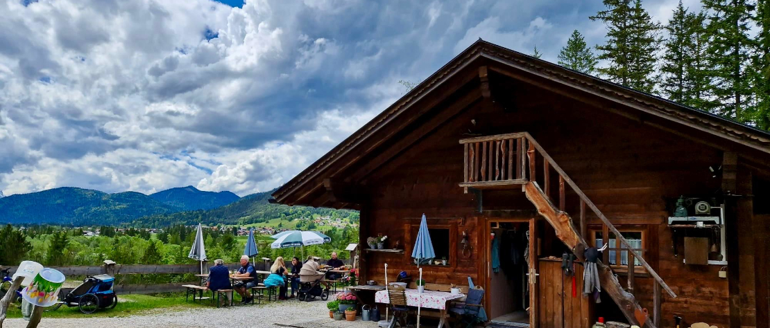Die Auhütte, eine kleine Holzhütte mit einem Tisch davor, recht Ausblick auf die Hügel und Berglandschaft, Blauer Himmel mit Wolken durchzogen